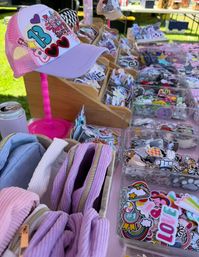 Pastel purple-pink trucker cap on a pink stand at an outdoor craft-fair vendor table, surrounded by colorful embroidered patches, stickers and ribbed pastel zip pouches