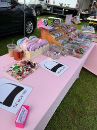 Outdoor craft market booth with a pink tablecloth showcasing trays of colorful patches, pins and charms, pastel scrunchies, decorated caps and 'Design Me!' hat templates for DIY custom hats.