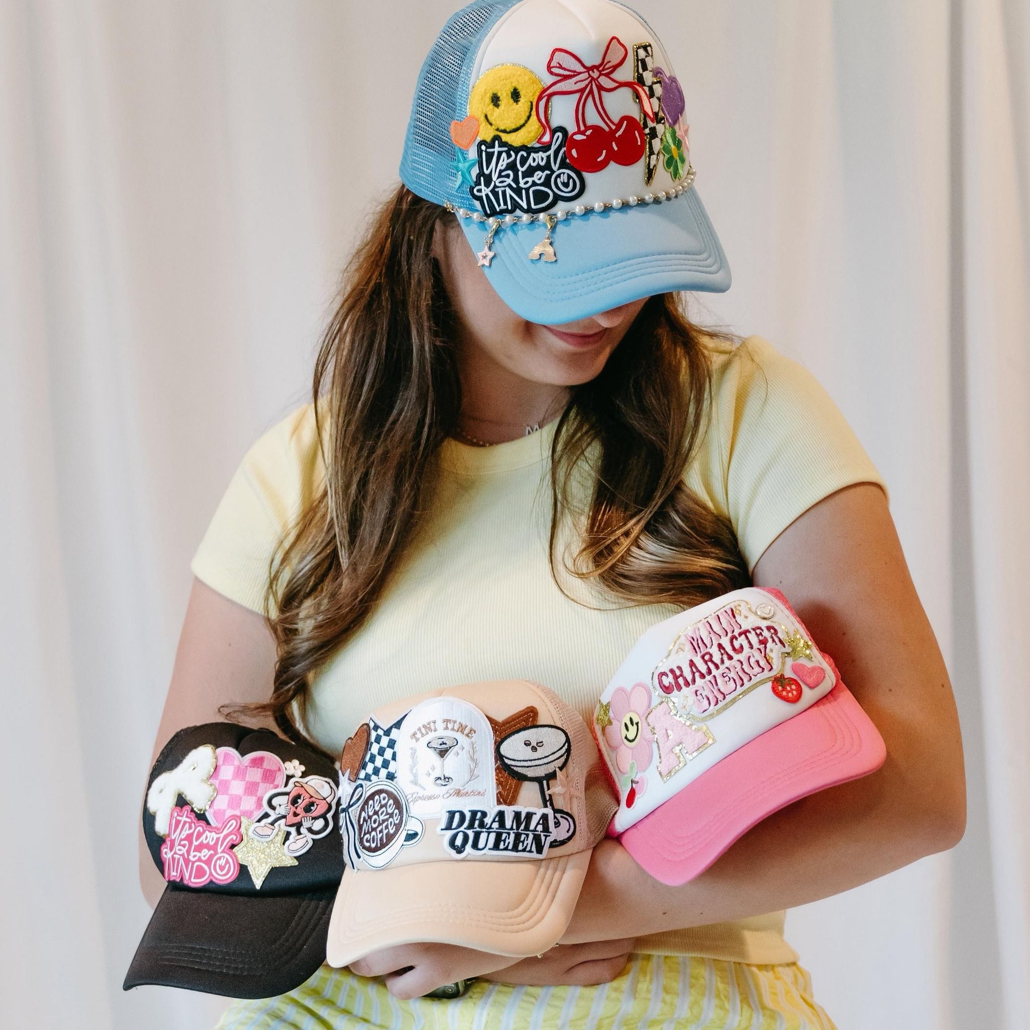 Person in yellow tee holding four playful embroidered trucker hats — blue cherry-and-smiley cap, black patchwork cap, beige Drama Queen cap, and pink-and-white character cap.
