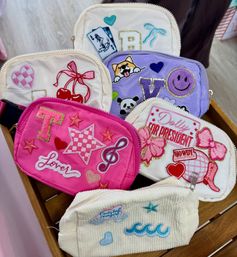 Bright pink, purple and cream embroidered zipper pouches with playful patches — cherries, checkered star, musical note, corgi, panda, smiley and 'Dolly for President' — arranged on a wooden display tray, cute kids' accessory and gift idea.