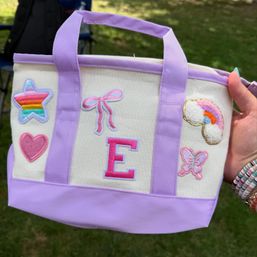 Lavender-trim canvas tote bag held by a hand in a grassy park, decorated with colorful patches including a rainbow star, pink heart, pink bow, bold letter E, fluffy rainbow cloud and pink butterfly.