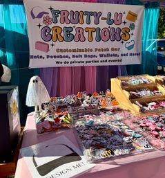 Colorful outdoor craft market booth with a playful pink‑and‑purple backdrop and large banner showcasing a customizable patch bar — clear trays and wooden bins overflowing with embroidered patches, stickers, pouches and a small ghost prop on a pink table.