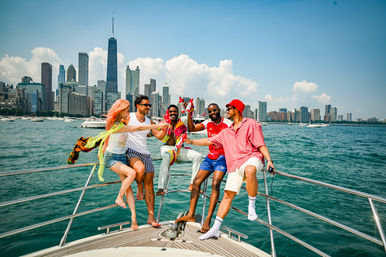 Five friends laughing and clinking bottles on a yacht in bright summer sun, turquoise water with boats and the Chicago skyline of skyscrapers in the background.