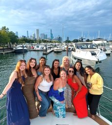 Ten friends smiling and posing on a yacht at a busy Chicago marina, with sailboats and the city skyline in the background under a partly cloudy sky.