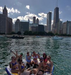 Group of friends in swimsuits on an inflatable raft in Lake Michigan, raising drinks with downtown Chicago skyline and boats along the shoreline on a sunny day