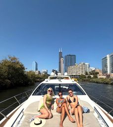 Three women relaxing on the bow of a white yacht cruising the Chicago River, with the Chicago skyline and Willis Tower rising against a clear blue sky.
