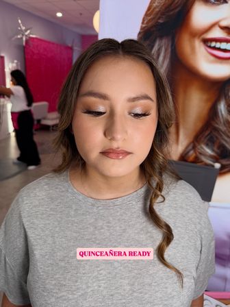 Teen girl in a beauty salon with quinceañera-ready glam: shimmery neutral eyeshadow, long lashes, glossy lips and a soft curled strand over a gray shirt with a “Quinceañera Ready” sticker
