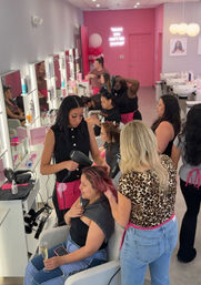 Busy pink-themed blowout bar hair salon interior with stylists blow-drying and styling multiple women at mirrored stations, sinks, neon wall sign and a client holding a champagne glass.