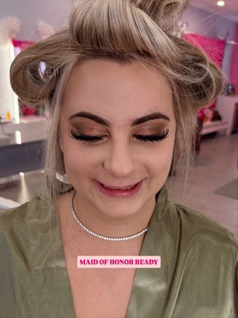 Close-up of a smiling maid of honor during bridal prep: blonde hair in rollers, shimmery bronze eyeshadow, dramatic false lashes, subtle pink lips, rhinestone choker and green satin robe in a salon setting.