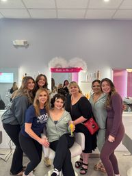Bridal party getting ready in a bright beauty salon — bride-to-be in a hoodie sits in a chair holding a mimosa, smiling friends gathered around under a pink “Bride to Be” sign.