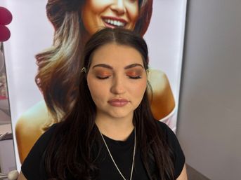 Young woman with eyes closed showing copper shimmer eyeshadow, glossy mauve lips and dark hair pinned back, posing in front of a large makeup display poster