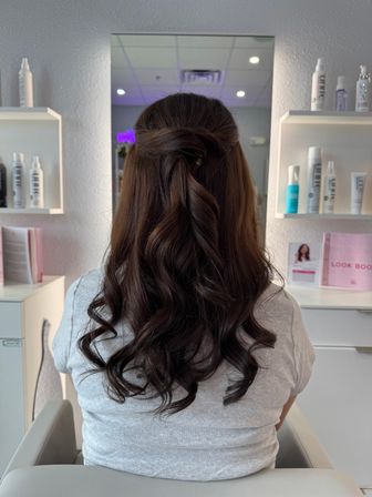 Back view of glossy brunette hair styled in a half-up twisted knot with loose cascading curls, seated in a modern salon chair with mirror and hair-product shelves in the background.
