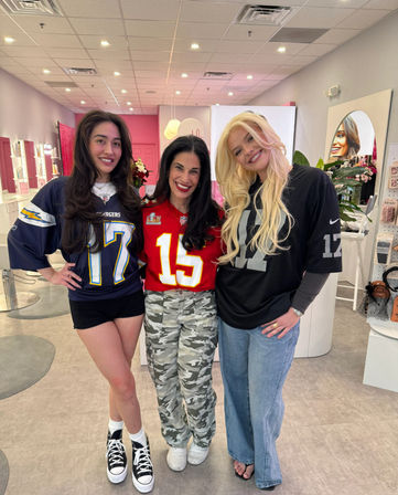 Three smiling women posing inside a pink beauty salon, each wearing football jerseys (navy #17, red #15, black #17) with casual shorts, camo pants, and jeans amid mirrors and product displays.