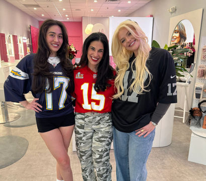 Three smiling women posing inside a pink beauty salon, each wearing football jerseys (navy #17, red #15, black #17) with casual shorts, camo pants, and jeans amid mirrors and product displays.