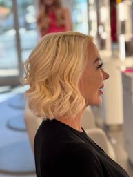 Side profile of a person with a chic shoulder-length blonde wavy bob haircut, soft textured waves styled in a bright modern hair salon