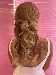 Half-up romantic twisted bun with loose blonde balayage curls cascading over a white dress, compass shoulder tattoo visible against a pink backdrop.