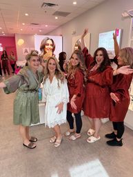 Bridal party getting ready in a pink-accented beauty salon — bride in a white lace robe smiling with friends in matching satin robes