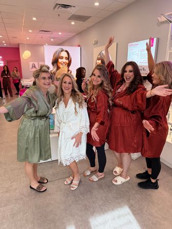 Bridal party getting ready in a pink-accented beauty salon — bride in a white lace robe smiling with friends in matching satin robes