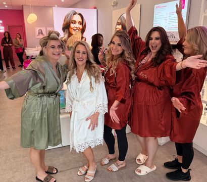 Bridal party getting ready in a pink-accented beauty salon — bride in a white lace robe smiling with friends in matching satin robes
