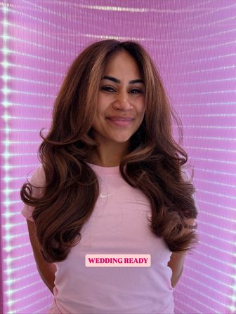 Smiling woman with long, voluminous wedding-ready waves and layers, wearing a pink tee against a bright pink LED backdrop, bridal hairstyle portrait.