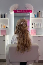 Romantic half-up bridal hairstyle with long loose waves and a crystal hairpiece, seated in a salon chair under a pink "Bride to Be" banner with styling products on nearby shelves.