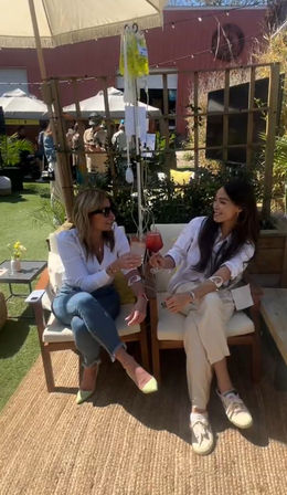 Two women smiling and toasting colorful drinks on a sunny outdoor patio lounge with umbrella, greenery and an IV drip stand in an urban courtyard setting