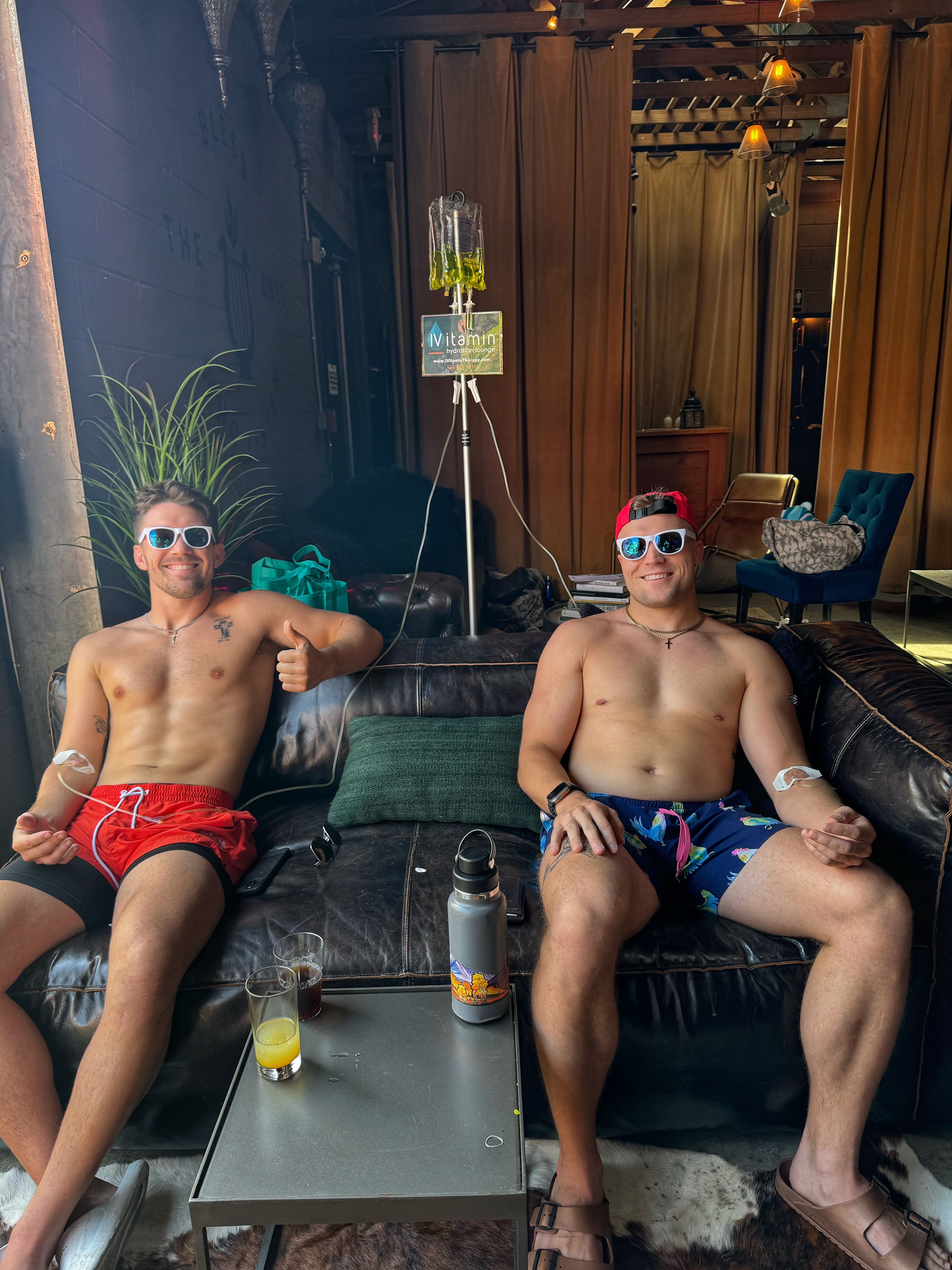 Two shirtless men in sunglasses and swim trunks reclining on a leather couch in a spa-like IV therapy lounge receiving vitamin drips, drinks and water bottle on a low table