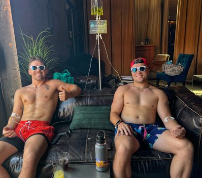 Two shirtless men in sunglasses and swim trunks reclining on a leather couch in a spa-like IV therapy lounge receiving vitamin drips, drinks and water bottle on a low table