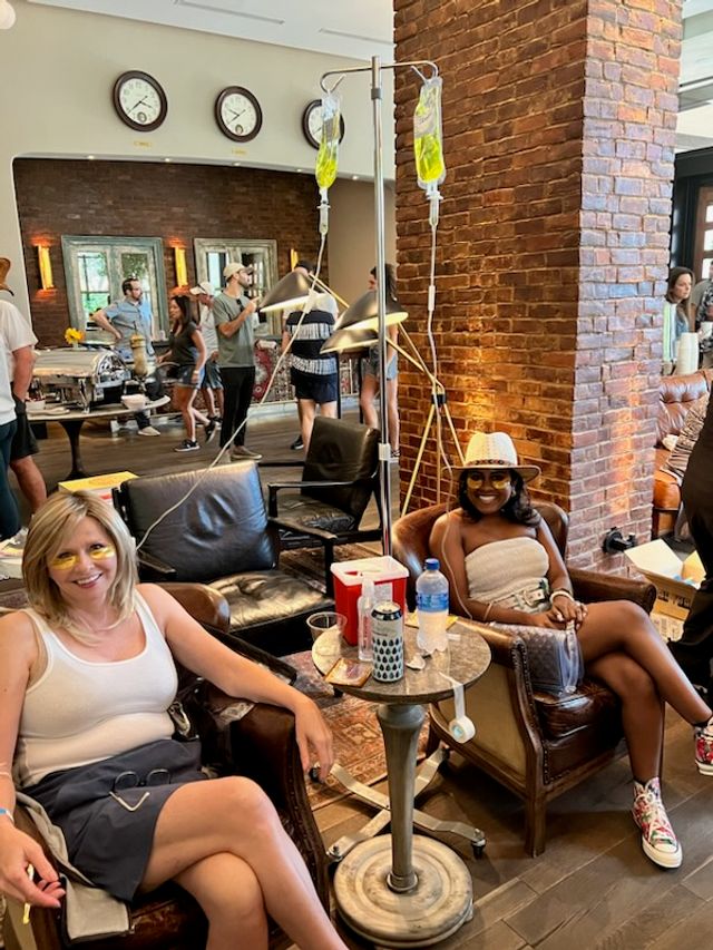 Two smiling women relax in leather armchairs in a cozy brick-walled lounge receiving yellow vitamin IV drips from hanging bags, with a small table of drinks, people mingling and a buffet area with clocks on the wall in the background.