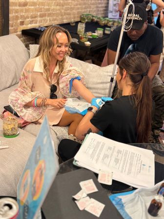 Smiling woman on a couch getting a blood draw from a gloved technician in a cozy brick-walled wellness pop-up, medical supplies and paperwork nearby.