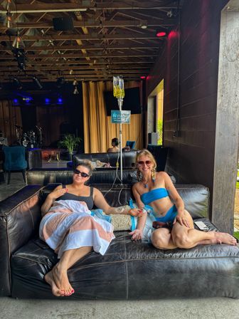 Two friends in swimwear lounging on a black leather sofa under a wooden-beamed patio, smiling while receiving IV vitamin drips at an outdoor wellness lounge.