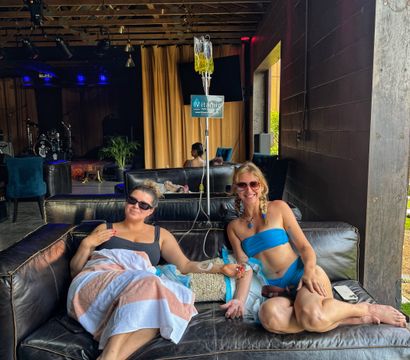 Two friends in swimwear lounging on a black leather sofa under a wooden-beamed patio, smiling while receiving IV vitamin drips at an outdoor wellness lounge.