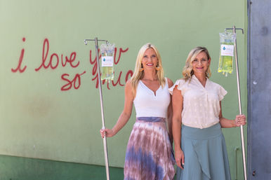 Two smiling women pose with IV hydration poles and yellow vitamin bags in front of a green wall mural that reads "i love you so much", wellness and IV therapy concept.