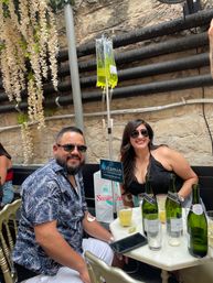 Two smiling people at a sunny urban patio table with wine bottles and cocktails, a bright yellow vitamin IV drip hanging from a pole, stone wall backdrop and trailing white flowers.