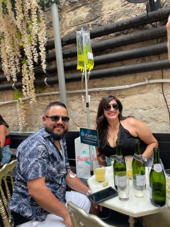 Two smiling people at a sunny urban patio table with wine bottles and cocktails, a bright yellow vitamin IV drip hanging from a pole, stone wall backdrop and trailing white flowers.