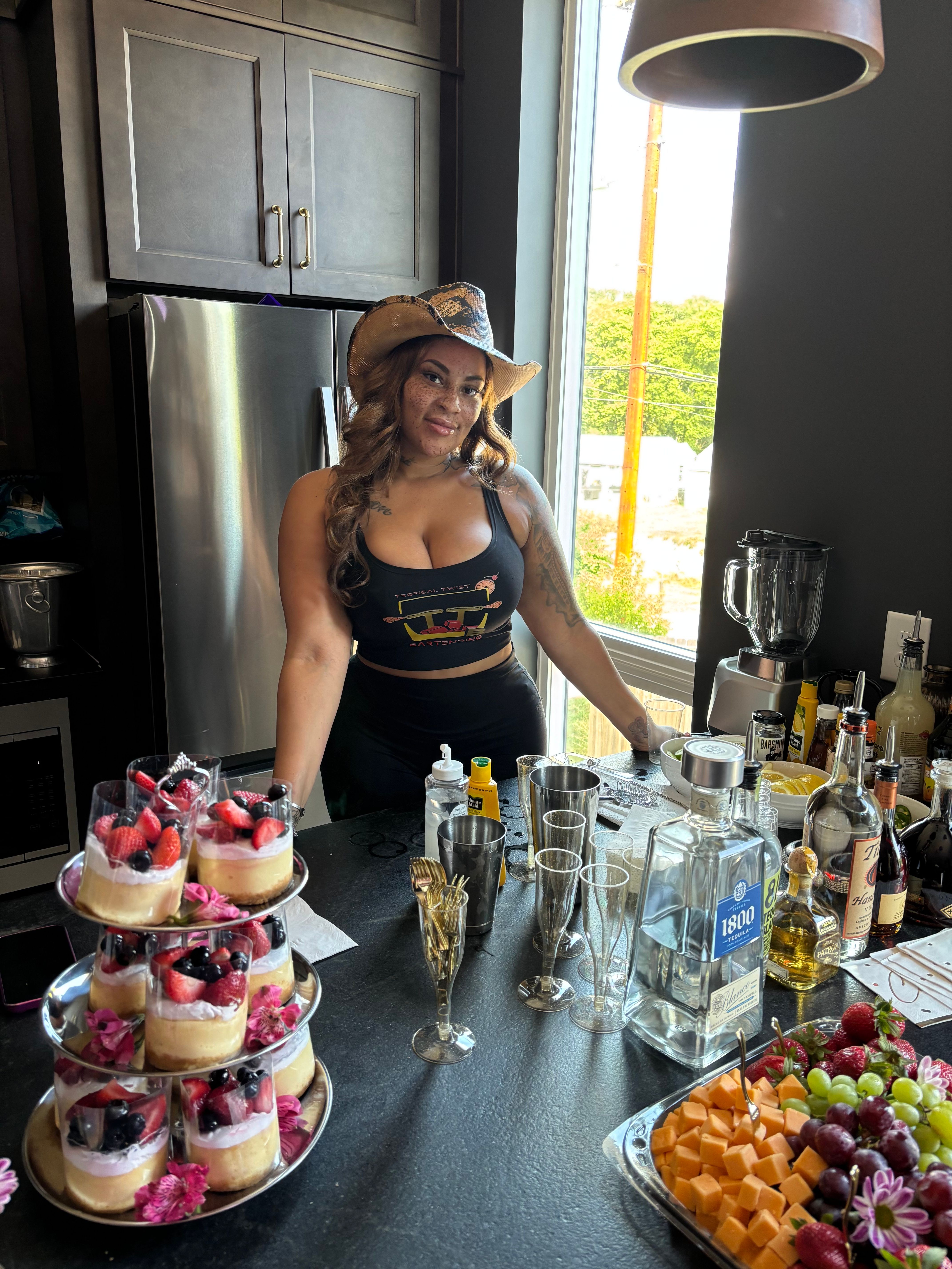 Woman in cowboy hat at a sunlit home kitchen bar with a dessert tower, cocktail tools and liquor bottles, plus a cheese-and-fruit platter for a party