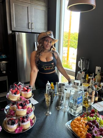 Woman in cowboy hat at a sunlit home kitchen bar with a dessert tower, cocktail tools and liquor bottles, plus a cheese-and-fruit platter for a party
