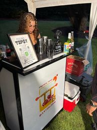 Outdoor backyard tented mobile bar at an evening event — bartender preparing drinks behind a white tropical-themed portable bar with a tip jar, QR-code payment sign, cocktail tools and bottled beers on the counter