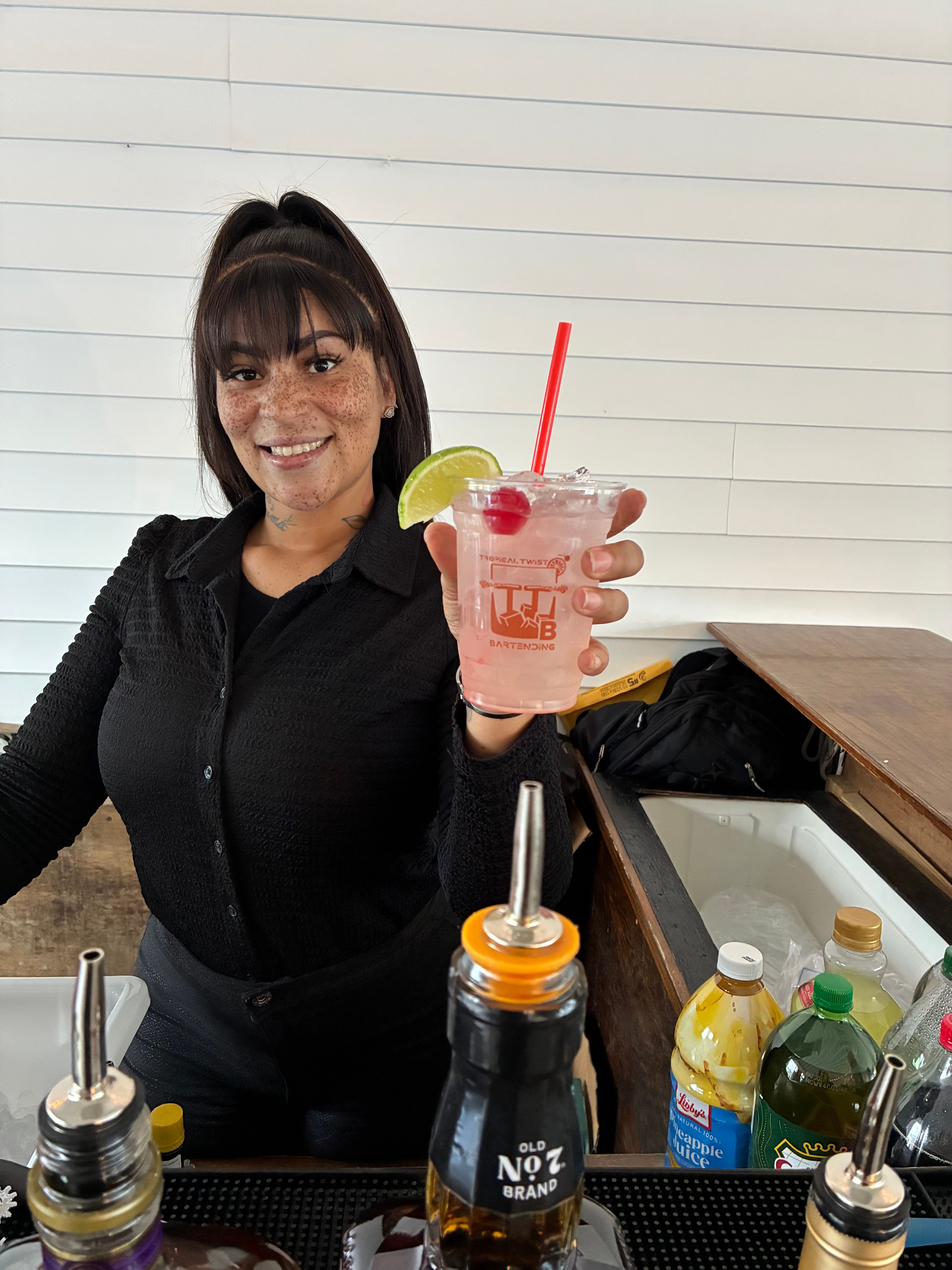 Smiling bartender behind a bar holding a pink iced cocktail in a plastic cup garnished with a lime wedge and cherry and a red straw, with bottles, mixers and an ice-filled service area in the foreground against a white shiplap wall.