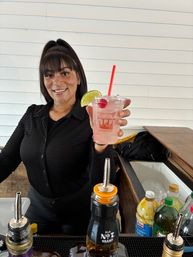 Smiling bartender behind a bar holding a pink iced cocktail in a plastic cup garnished with a lime wedge and cherry and a red straw, with bottles, mixers and an ice-filled service area in the foreground against a white shiplap wall.