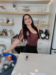 Woman with long dark hair and glasses posing in a modern white kitchen, holding a sparkling wine bottle beside a blue ice bucket of beers on a marble island with open wood shelves and dishes in the background.