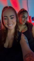 Two smiling friends taking a selfie in a neon-lit bar with red and blue lighting, wearing black tops with visible tattoos and jewelry