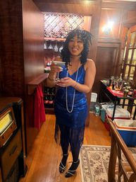 Person in blue sequin 1920s flapper dress and pearls holding a cocktail in a cozy wood-paneled home bar with wine rack and hardwood floors.