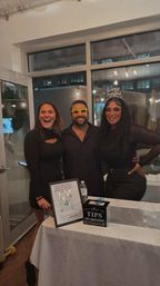 Three friends in black outfits smiling behind a table with a tip box and QR sign at an indoor New Year’s celebration — man wearing yellow novelty glasses and a woman with a “Happy New Year” headband in a loft-style venue with string lights.