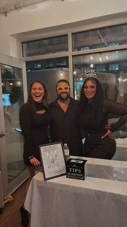 Three friends in black outfits smiling behind a table with a tip box and QR sign at an indoor New Year’s celebration — man wearing yellow novelty glasses and a woman with a “Happy New Year” headband in a loft-style venue with string lights.