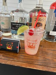 Pink tropical cocktail in a clear plastic cup with crushed ice, garnished with a lime wedge and red cherry, sitting on a wooden bar counter with clear liquor bottles and bartending cards in the background.