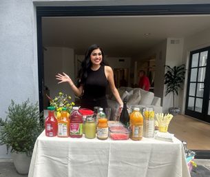 Smiling woman standing behind a DIY beverage bar at a modern open-plan home, table topped with colorful juice bottles, mason jars, sliced watermelon, glasses and yellow-striped straws, with potted plants and a sofa visible indoors.