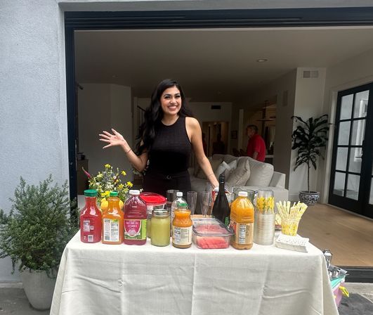 Smiling woman standing behind a DIY beverage bar at a modern open-plan home, table topped with colorful juice bottles, mason jars, sliced watermelon, glasses and yellow-striped straws, with potted plants and a sofa visible indoors.
