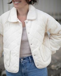 Cropped image of a smiling woman in a cream quilted jacket, beige knit top, silver necklace and high‑waisted blue jeans — casual street style