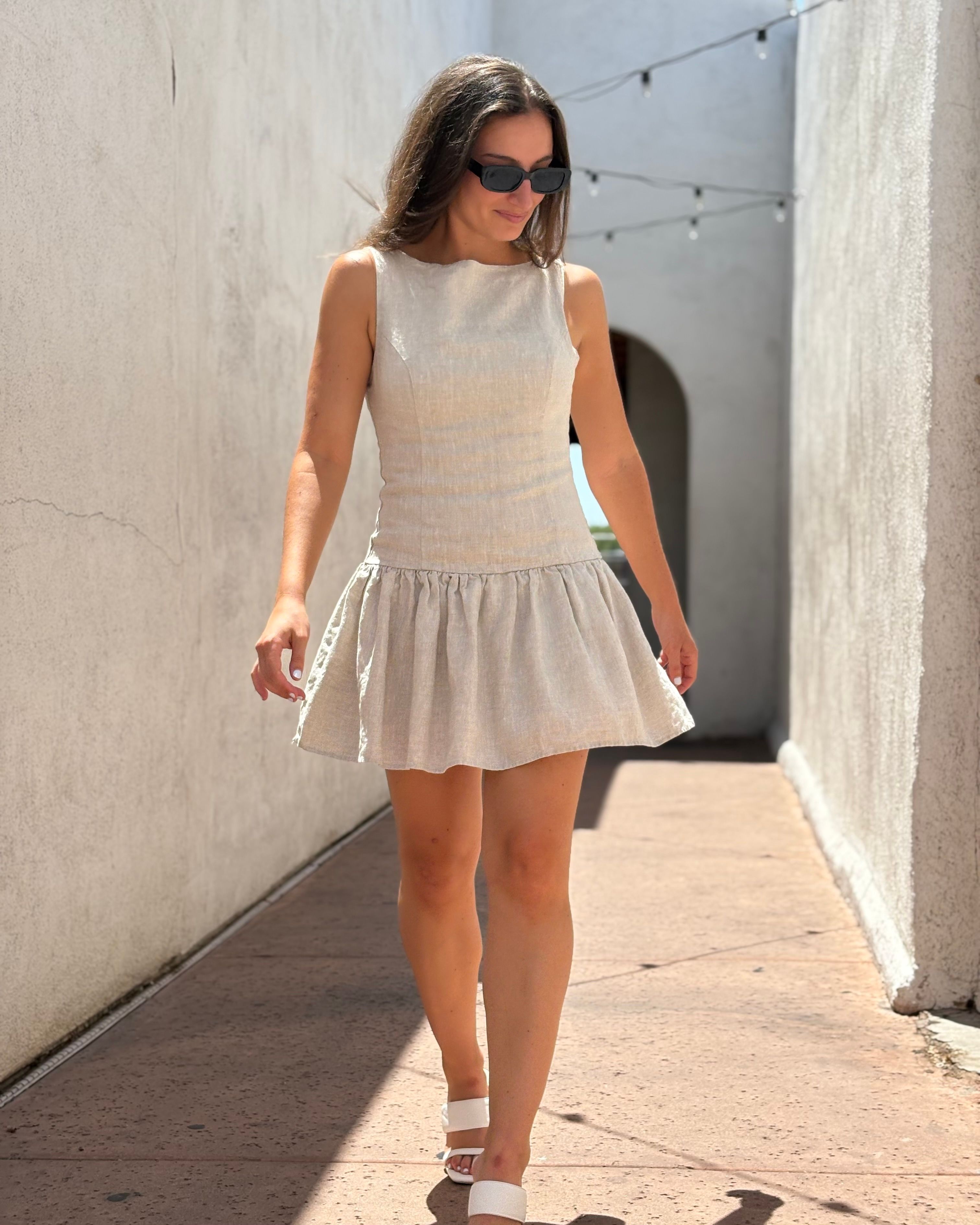 Woman in a sleeveless beige linen sundress, white sandals and black sunglasses walking down a sunlit stucco alley with string lights — summer street style.